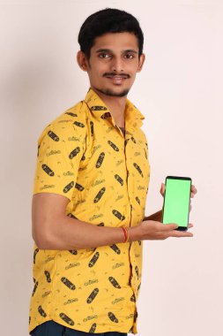 Tıraşsız, çekici, sarı tişörtlü, kamerada cep telefonu gösteren ve el işaretli, beyaz arka plandaki stüdyo resminde izole edilmiş Hintli bir adam.