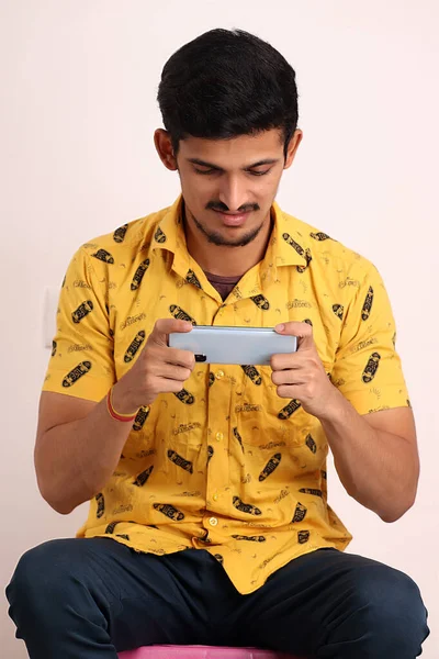 穿着黄色衬衫 拿着机器人智能手机玩游戏 脸上挂着白色背景图片 坐在椅子上 摆出一副喜怒无常的样子 没有刮胡子的迷人印度男人 — 图库照片