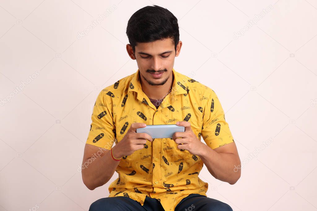 unshaven attractive Indian guy with wearing yellow shirt and Playing games on android smartphone on and gesturing emotion sign with face isolated on white background studio portrait & seating on chair