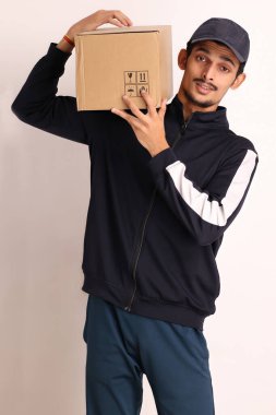 Kahverengi kutulu, siyah tişörtlü, beyaz arka planda yüz ifadeleri olan, traşsız Hintli kurye çocuk. 