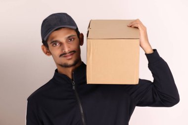 Kahverengi kutulu, siyah tişörtlü, beyaz arka planda yüz ifadeleri olan, traşsız Hintli kurye çocuk. 