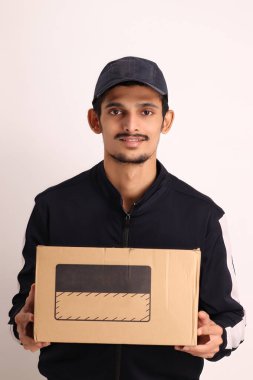 Kahverengi kutulu, siyah tişörtlü, beyaz arka planda yüz ifadeleri olan, traşsız Hintli kurye çocuk. 