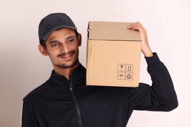 Kahverengi kutulu, siyah tişörtlü, beyaz arka planda yüz ifadeleri olan, traşsız Hintli kurye çocuk. 