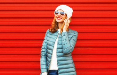 Akıllı telefondan konuşan mutlu genç bir kadının portresi. Ceket ve beyaz şapkalı, kırmızı arka planda ceketli.