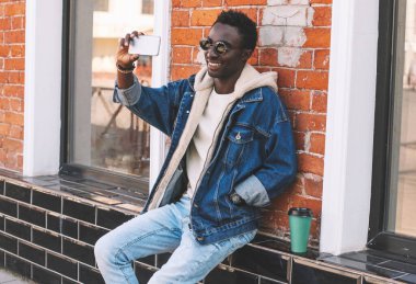 Mutlu gülümseyen Afrikalı genç bir adamın portresi. Şehir caddesinde akıllı telefonla selfie çekiyor.