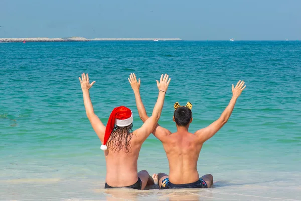 Dos hombres en trajes de baño y sombreros de Santa Claus y Tigre están sentados en la playa — Foto de Stock