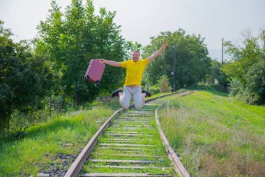 Kırmızı bavullu şortlu bir adam demiryoluna atlıyor.
