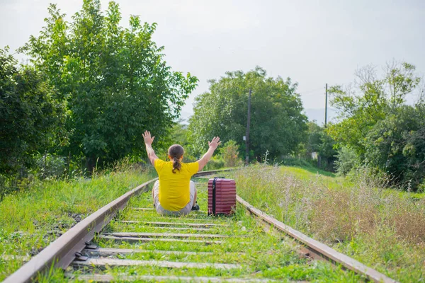 Cheerful traveler with a red suitcase on the railroad sits — Stock Photo, Image