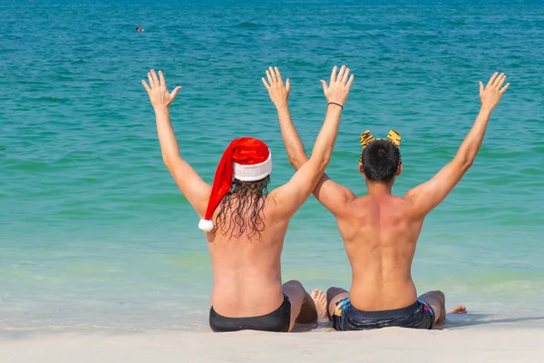 수영복을 입고 산타클로스와 호랑이 모자를 쓴 두 남자 가 해변에 앉아 있습니다 — 스톡 사진