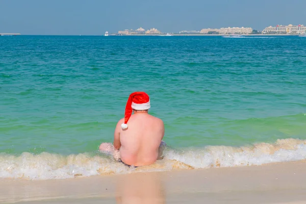 해변에 앉아 있는 뚱뚱 한 남자는 신년 모자를 쓰고 있다 — 스톡 사진