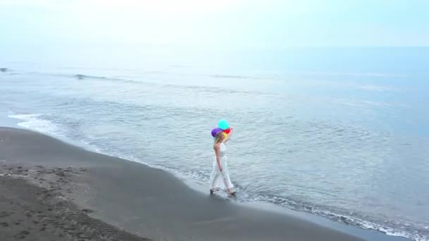 Seorang gadis dengan gaun putih dengan bola berwarna berjalan di pantai dengan anjing — Stok Video