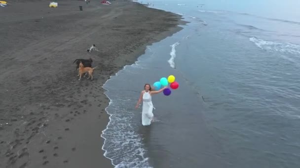 Jedna dziewczyna w białej sukience z kolorowymi kulkami biegnie po plaży z psami — Wideo stockowe