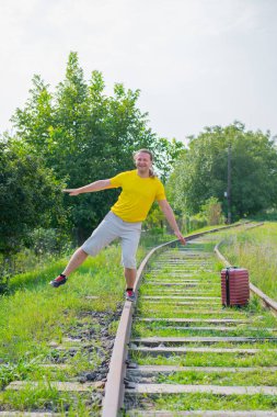 Uzun saçlı esmer gezgin, elinde bavuluyla raylarda yürüyor.