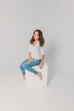 Beyaz gömlekli ve kotlu bir kız beyaz arka planda beyaz bir küpün üzerinde oturuyor.