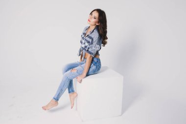 Ekose gömlekli ve kotlu bir kız beyaz arka planda beyaz bir küpün üzerinde oturuyor.