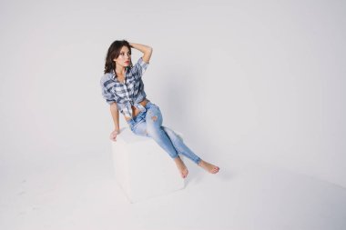 Ekose gömlekli ve kotlu bir kız beyaz arka planda beyaz bir küpün üzerinde oturuyor.