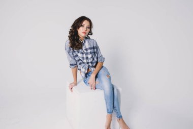 Ekose gömlekli ve kotlu bir kız beyaz arka planda beyaz bir küpün üzerinde oturuyor.