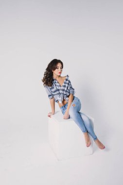 Ekose gömlekli ve kotlu bir kız beyaz arka planda beyaz bir küpün üzerinde oturuyor.