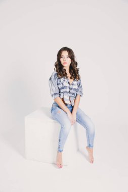 Ekose gömlekli ve kotlu bir kız beyaz arka planda beyaz bir küpün üzerinde oturuyor.