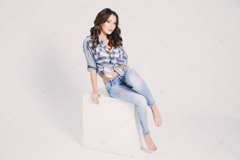 a girl in a plaid shirt and jeans sits on a white cube on a white background