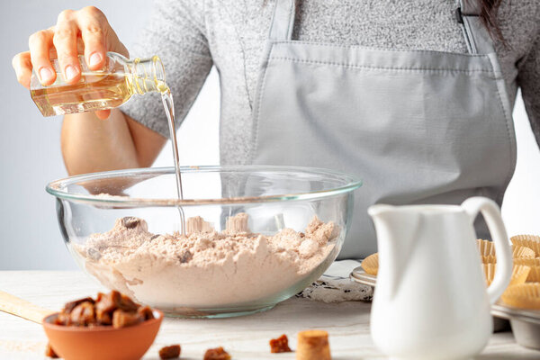 A woman is adding ingredients for making delicious fruit and nut cupcake on white marble countertop background. Muffin tin with liner, ingredients, and utensils are seen. Caucasian woman wearing apron