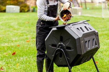 Bir kadın arka bahçedeki açık havada takla atan bir beze küçük bir mutfak artıklarını atıyor. Bu metal bacaklı plastik birimler daha iyi havalandırma ve hızlı gübreleme için geri dönebilirler..