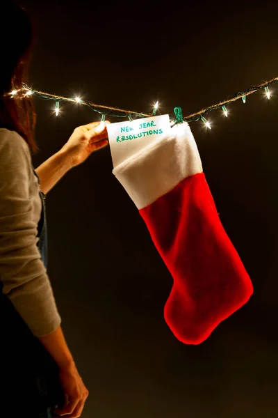 A woman is hanging a notice saying new year resolutions to a a gift stocking on a string of decorative lights. A quirky dark humor concept image for a reminder for new changes in lifestyle.