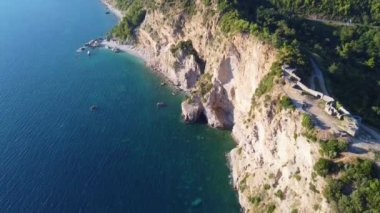 Mogren Kalesi 'nin hava manzarası turkuaz deniz ile parlak güneş ışığı altındaki yeşil sık ormanlar arasındaki harabeleri gösteriyor, Budva, Karadağ