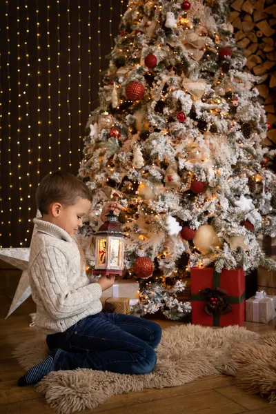 Petit garçon tenant une lanterne blanche dans sa main et assis près du sapin de Noël à la maison, Bonne année 2022 — Photo