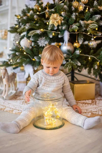 Adorable petite fille assise près du sapin de Noël avec des lumières festives et des cadeaux de Noël. Noël et Nouvel An. — Photo