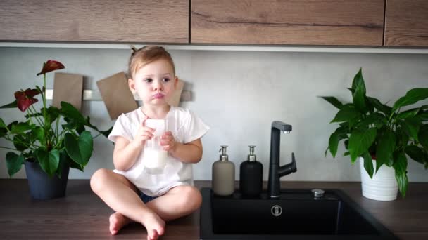 Niña feliz sentada en la mesa en la cocina y bebiendo leche — Vídeos de Stock