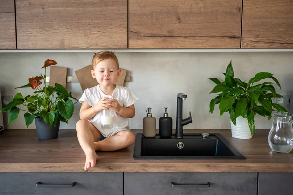 Boldog kislány ül az asztalnál a konyhában és tejet iszik. — Stock Fotó