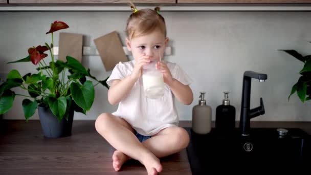 Glückliches kleines Mädchen sitzt am Tisch in der Küche und trinkt Milch — Stockvideo