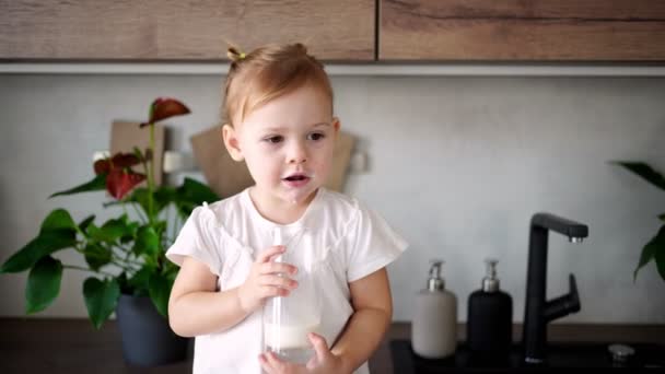 Glad liten flicka sitter vid bordet i köket och dricker mjölk — Stockvideo