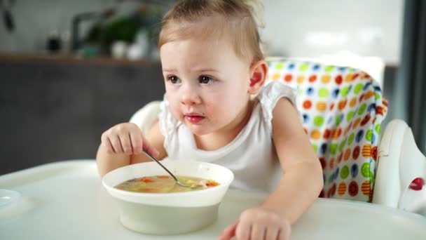 Cute baby girl maluch siedzi na wysokim krześle i jedzenie jej zupy lunchowej w domu kuchnia. — Wideo stockowe