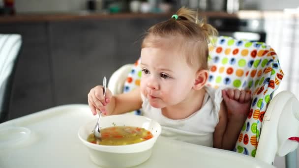 可爱的小女孩蹒跚学步，坐在高高的椅子上，在家里厨房里吃午饭汤. — 图库视频影像