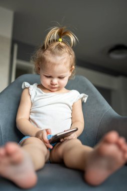 Gülümseyen küçük kız koltukta oturuyor ve akıllı telefon kullanıyor.