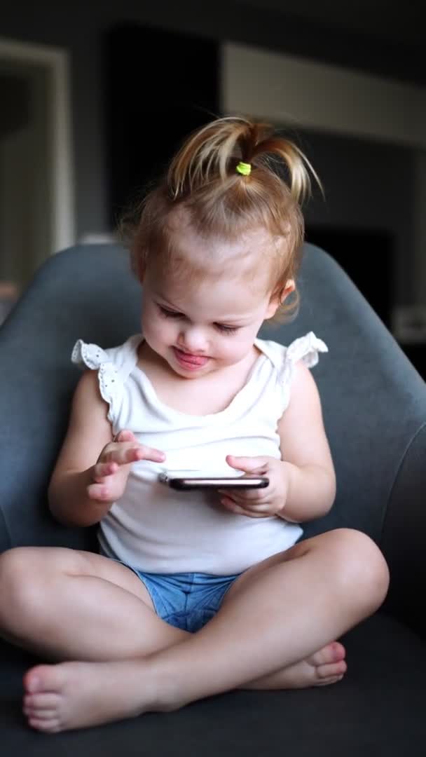 La niña sonriente está sentada en un sillón y usando un teléfono inteligente — Vídeos de Stock
