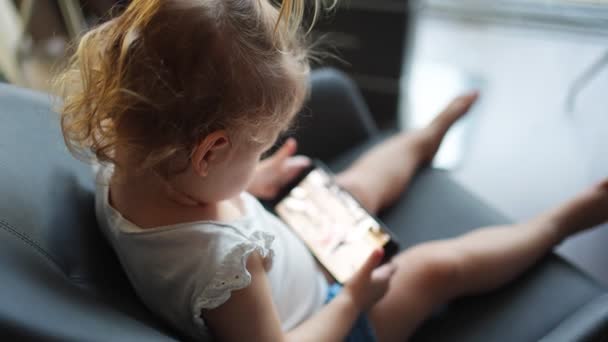 La niña sonriente está sentada en un sillón y usando un teléfono inteligente — Vídeos de Stock