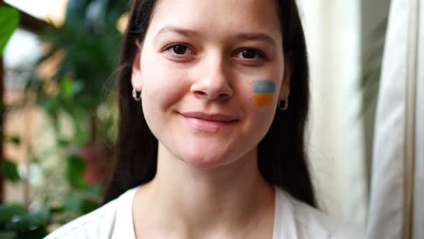 Усміхнена молода українська дівчина з прапором України на обличчі дивиться на камеру. Концепція участі українського народу у війні з Росією. Не військова концепція — стокове відео