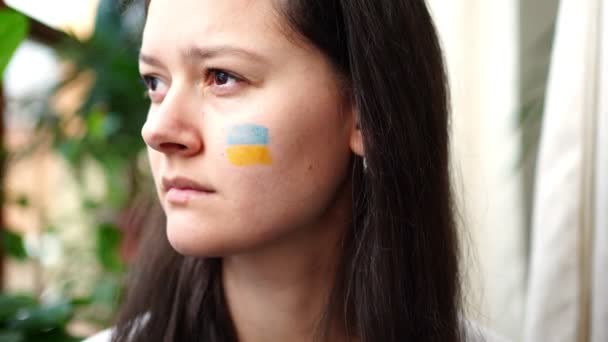 Μια λυπημένη νεαρή Ουκρανή με τη σημαία της Ουκρανίας στο πρόσωπό της κοιτάζει μέσα από το παράθυρο. Η έννοια της συμμετοχής του ουκρανικού λαού στον πόλεμο με τη Ρωσία. Όχι πολεμική έννοια — Αρχείο Βίντεο