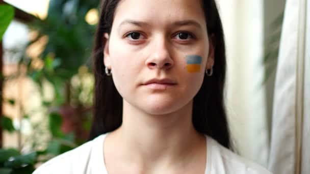 Une jeune fille ukrainienne triste avec le drapeau de l'Ukraine sur son visage regarde vers la caméra. Le concept de participation du peuple ukrainien à la guerre contre la Russie. Pas concept de guerre — Video