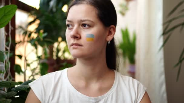 Ein trauriges junges ukrainisches Mädchen mit der Fahne der Ukraine auf dem Gesicht blickt durch das Fenster. Das Konzept der Beteiligung des ukrainischen Volkes am Krieg gegen Russland. Kein Kriegskonzept — Stockvideo