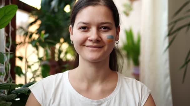 Une jeune fille ukrainienne souriante avec le drapeau de l'Ukraine sur son visage regarde vers la caméra. Le concept de participation du peuple ukrainien à la guerre contre la Russie. Pas concept de guerre — Video