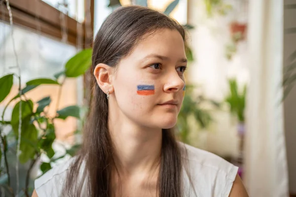 A sad young Russian girl with the flag of Ukraine on her face is looking through window. The concept of participation of the Ukrainian people in the war with Russia. Not war concept