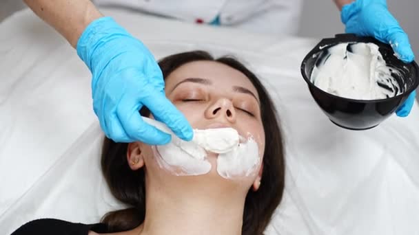 Läkare kosmetolog eller hudläkare gör ansiktsmask i kosmetologi salong. Professionell kosmetolog applicera ansiktsmask på kaukasisk kvinna ansikte liggande på sängen i badrock. — Stockvideo