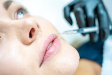 Kadının yüzünün ve elinin dudaklarının yakınında şırıngayı tutan ameliyat eldiveniyle güzellik tedavisine hazır bir şekilde çekilmiş fotoğrafı. Enjeksiyon kozmetolojisi, dudak büyütme ve düzeltme konsepti.