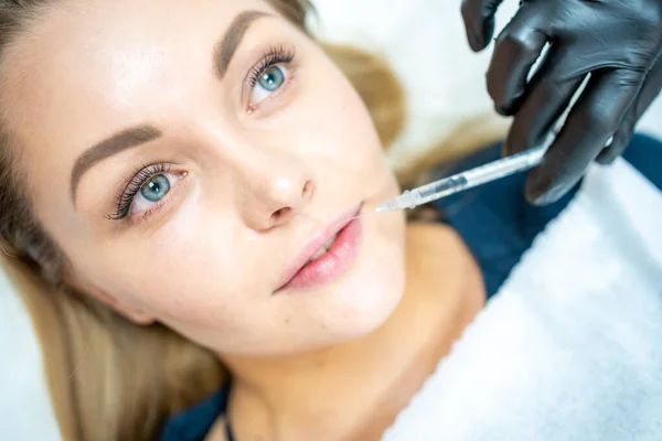 Close-up of woman face and hand in surgical glove holding syringe near her lips, ready to receive beauty treatment. Injection cosmetology, lips augmentation and correction concept.