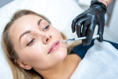 Kadının yüzünün ve elinin dudaklarının yakınında şırıngayı tutan ameliyat eldiveniyle güzellik tedavisine hazır bir şekilde çekilmiş fotoğrafı. Enjeksiyon kozmetoloji, estetik cerrahi, dudak büyütme ve düzeltme konsepti.