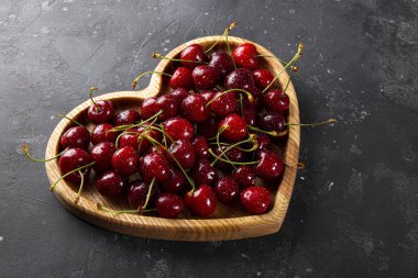 Olgun kiraz ve çilek ahşap bir tabakta, kalp şeklinde ve koyu gri arka planda, Sevgililer Günü kartında meyve var.